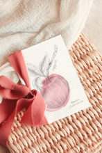 Load image into Gallery viewer, A small artwork featuring a watercolor sketch of an apple, tied with a coral pink ribbon and displayed on a woven wicker basket with a soft cream textile background.