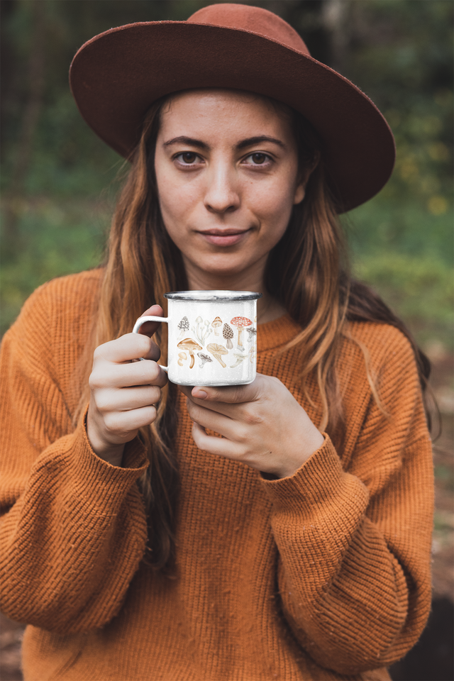 Lady in tan sweater and hate holding a small white and metal camper mug with mushroom printed around the mug. 