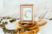 Load image into Gallery viewer, A framed botanical art print initail "C" featuring a Calla Lily with a small butterfly, styled on a yellow knit blanket with dried flowers and eucalyptus leaves on a bed.