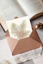 Load image into Gallery viewer, A small artwork with a watercolor illustration of oak leaves and an acorn, displayed inside a brown kraft paper envelope. The scene is styled with an open book, a gold acorn, and a white lace doily on a gray fabric background.
