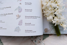 Load image into Gallery viewer, Open book with floral illustrations and text, accompanied by a bouquet of white flowers.
