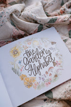 Load image into Gallery viewer, Book titled 'The Garden Alphabet' with floral designs on a floral fabric background