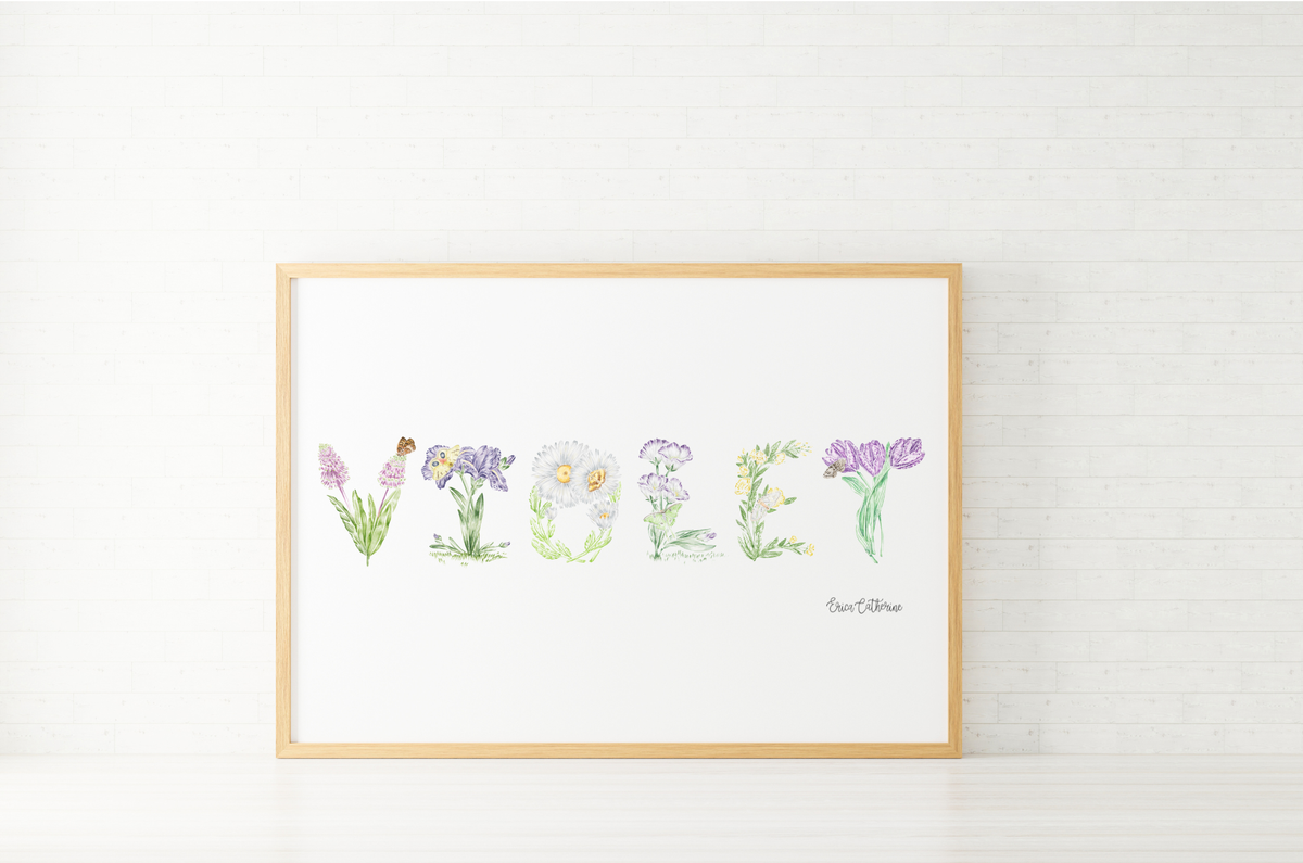 A framed watercolor art print featuring the word 'Violet' spelled out with delicate botanical illustrations, set in a light wood frame against a white brick wall background.