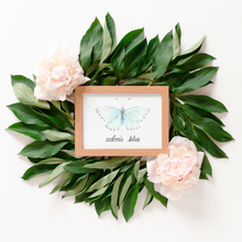 Load image into Gallery viewer, The framed butterfly print is of the Adonis blue butterfly, a light blue butterfly. The Print is framed in a light warm wood frame. Large green leaves and light pink flowers surround the framed print.

