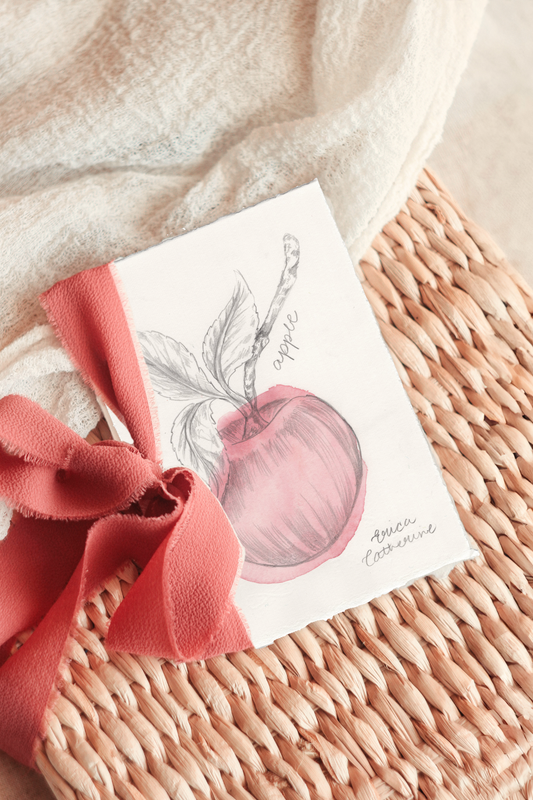 A small artwork featuring a watercolor sketch of an apple, tied with a coral pink ribbon and displayed on a woven wicker basket with a soft cream textile background.