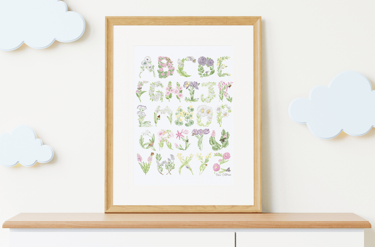 A framed alphabet art print featuring the alphabet letters designed as watercolor wildflowers, displayed on a light wood shelf against a cream wall with decorative cloud cutouts.