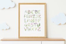 Load image into Gallery viewer, A framed alphabet art print featuring the alphabet letters designed as watercolor wildflowers, displayed on a light wood shelf against a cream wall with decorative cloud cutouts.
