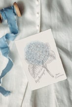 Load image into Gallery viewer, A small white art print featuring a delicate pencil-style illustration of a hydrangea bouquet with the handwritten text 'hydrangea' and 'Bree Calmmore,' styled on white fabric next to a blue ribbon.
