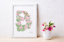 Load image into Gallery viewer, A framed botanical letter "B" art print featuring the a pink begonias and a moth. Styled on a light wood surface next to a white ceramic vase with pink flowers against a textured white wall.
