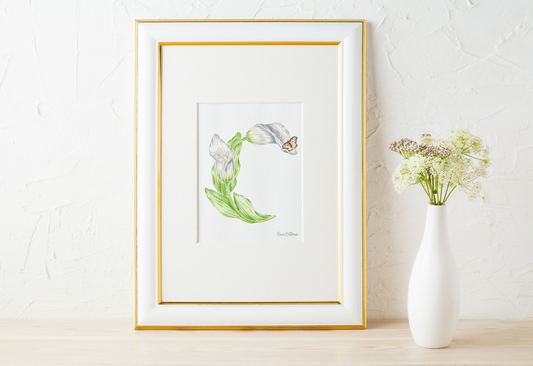 A framed botanical art print featuring a white Calla Lily and butterfly forming the letter C, styled on a light wood surface next to a white ceramic vase with flowers against a textured white wall.