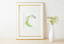 Load image into Gallery viewer, A framed botanical art print featuring a white Calla Lily and butterfly forming the letter C, styled on a light wood surface next to a white ceramic vase with flowers against a textured white wall.
