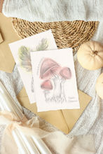 Load image into Gallery viewer, A styled flat-lay of two botanical artworks featuring mushroom illustrations, surrounded by kraft envelopes, a woven placemat, a soft gray blanket, and small pumpkins.
