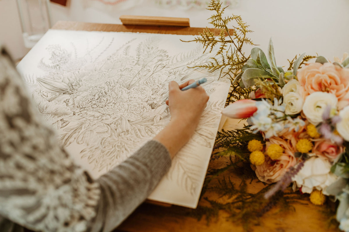 The artist Erica Catherine is sitting at a desk. She is drawing a floral bouquet in pencil and the bouquet she is referencing is sitting next to her drawing on the table.