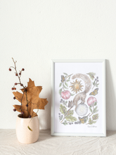 Load image into Gallery viewer, A framed botanical art print with a whimsical creature illustration, styled on a light fabric surface next to a small vase with dried leaves and berries against a textured white wall.
