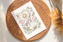 Load image into Gallery viewer, A botanical art print featuring a squirrel, sun, moon, and plants styled on a round woven placemat with dried pampas grass in the background.

