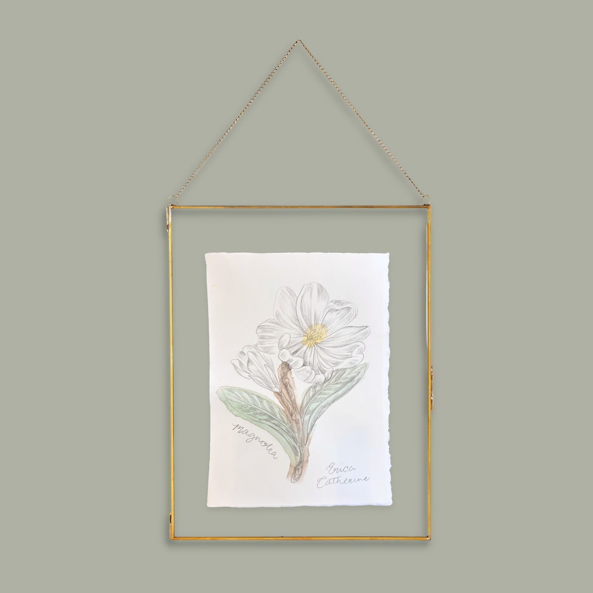 A framed botanical art print featuring a white flower with green leaves, hanging from a gold metal frame and chain against a solid sage green background.