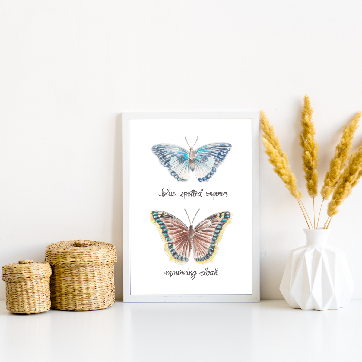 Framed art print in a white frame. To the left are two small whicker baskets, and to the right is a white geometric vase with fluffy wheat grass. The framed print is of two butterflies, one above the other. The top butterfly is called the Blue spotted emperor and is a blue and white butterfly. The lower butterfly is called the mourning cloak, it is a brown butterfly with blue spots around the edge and a yellow band around the outside.
